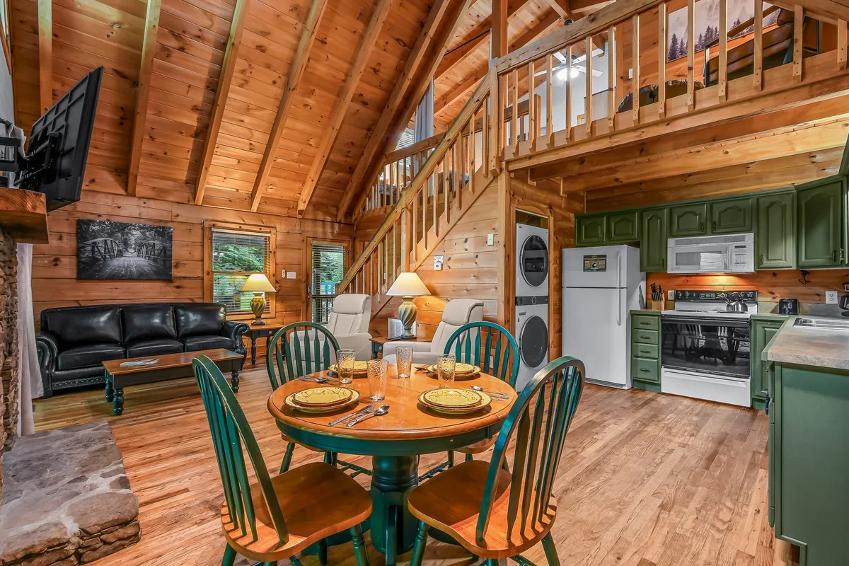 A spacious interior with a round dining table set with dishes and glasses, a sleek black leather sofa positioned near a stone fireplace, and an open staircase. The setup is ideal for family meals, board games, or relaxing by the fire with loved ones.
