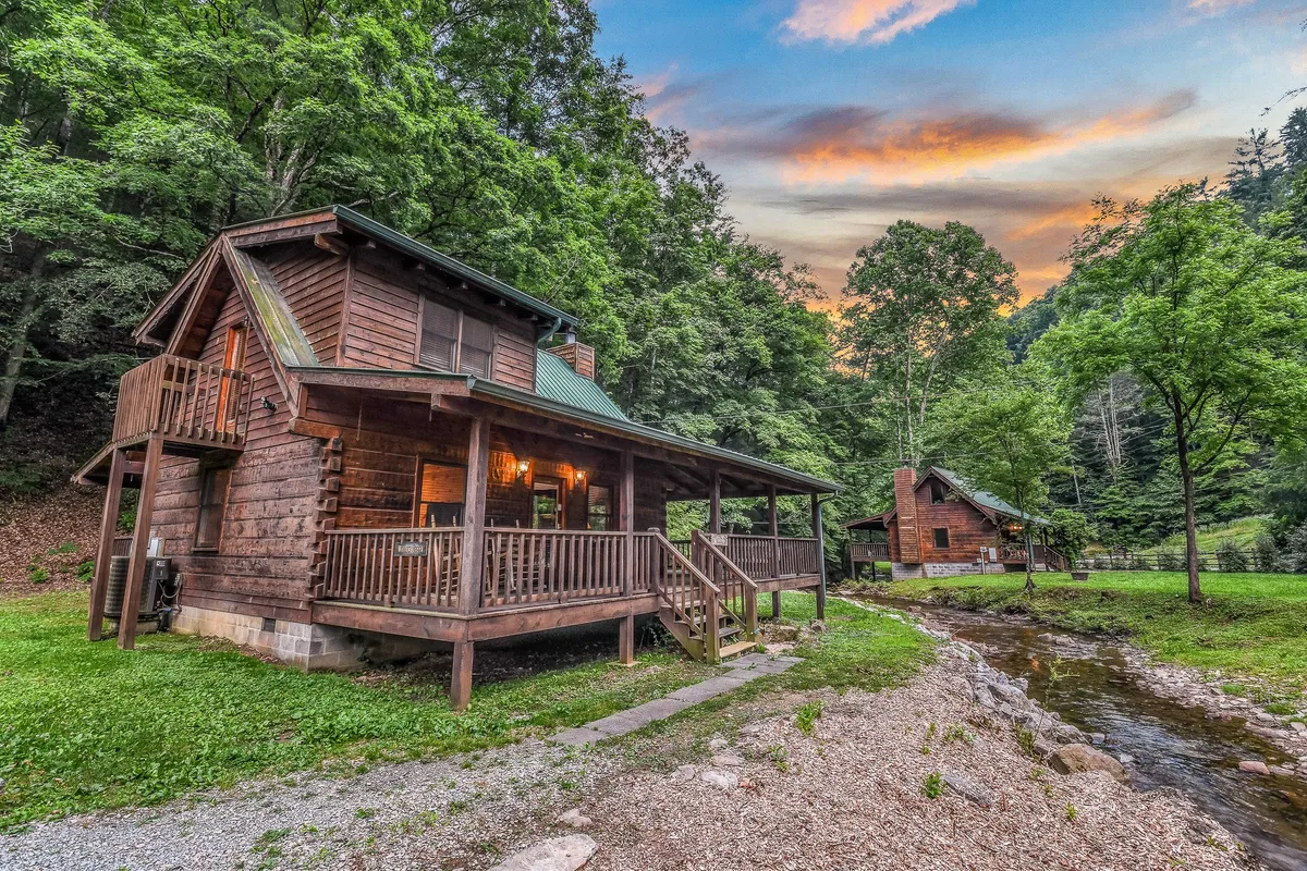 This wide-angle exterior photo captures the log cabin nestled beside a picturesque stream, framed by mature trees and a glowing sunset sky. The porch and stream are both prominent, inviting families to relax, picnic, or enjoy the natural beauty right at their doorstep. The tranquility of the scene evokes the peaceful, adventurous spirit described in the section.