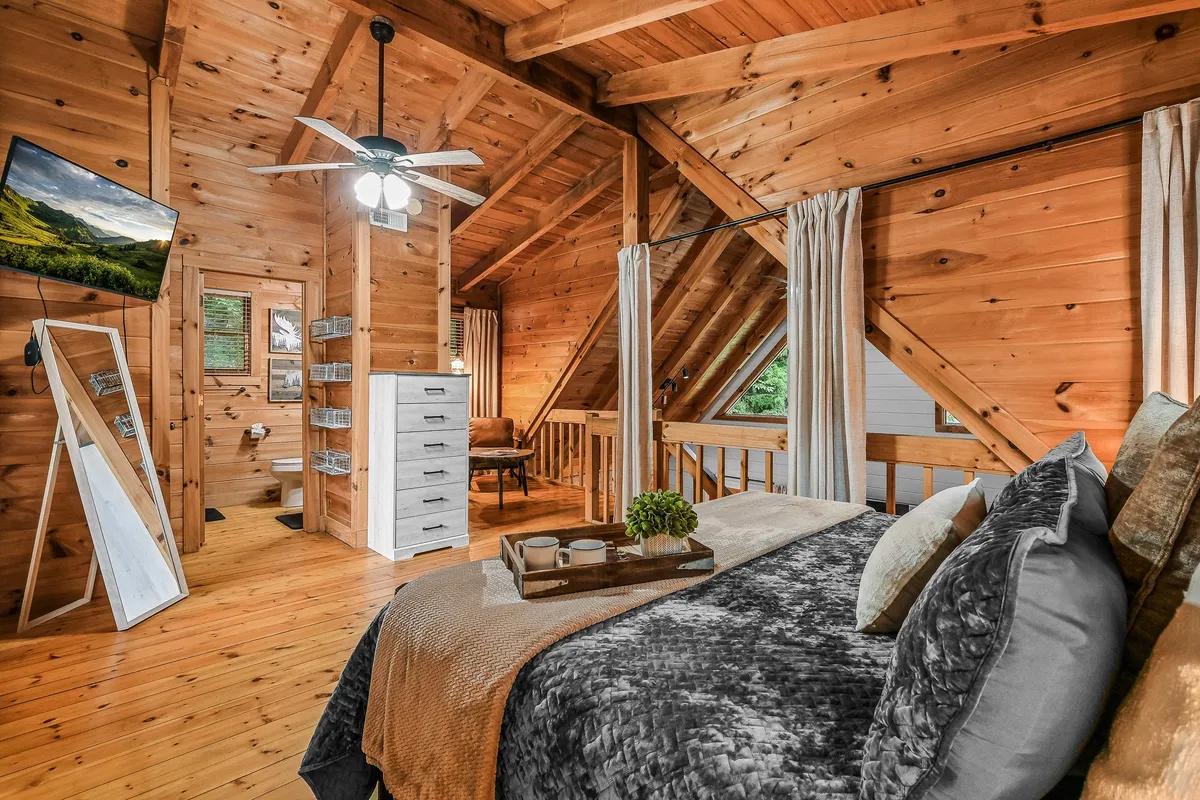 A large bed with textured bedding sits beneath exposed wooden ceiling beams, with natural light streaming in through windows. The room includes a mounted TV, a dressing mirror, and a comfortable chair, reinforcing a serene and luxurious environment.