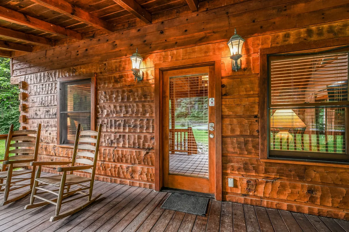 A charming porch scene featuring wooden rocking chairs and warm outdoor lighting flanking the cabin’s entrance. The inviting setup suggests slow mornings or starlit evenings spent enjoying the serene sounds of nature.