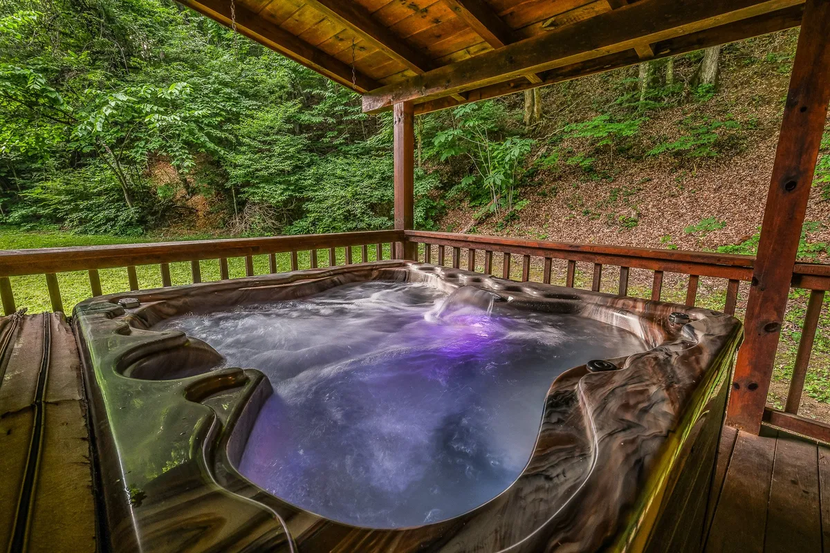 An exterior shot highlighting a bubbling hot tub tucked into a wooden structure, surrounded by lush greenery. The hot tub is positioned for privacy and relaxation, with nature in every direction. This setting is ideal for unwinding after a day of play or exploration, appealing to adults and families alike.