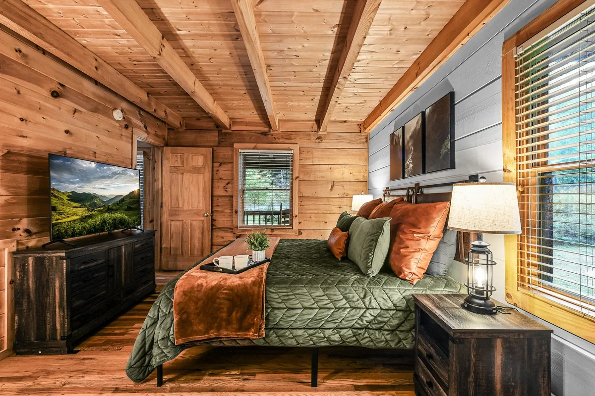 A spacious king-sized bed adorned with green and orange bedding sits at the center of a warmly lit bedroom. The room features wooden furniture, a TV showcasing a scenic view, and a bedside lamp that casts a cozy glow, creating an inviting and restful atmosphere.