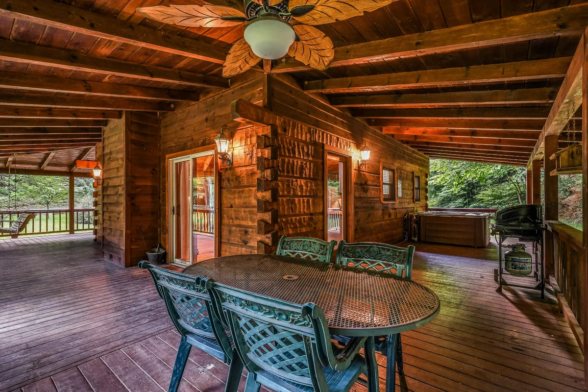 This image features a cozy dining area with a wooden table beneath ceiling fans and exposed beams, opening onto a deck area with a visible hot tub. The scene invites guests to dine, socialize, and relax while enjoying a seamless transition between indoor comfort and outdoor serenity, with hints of greenery visible through the windows.