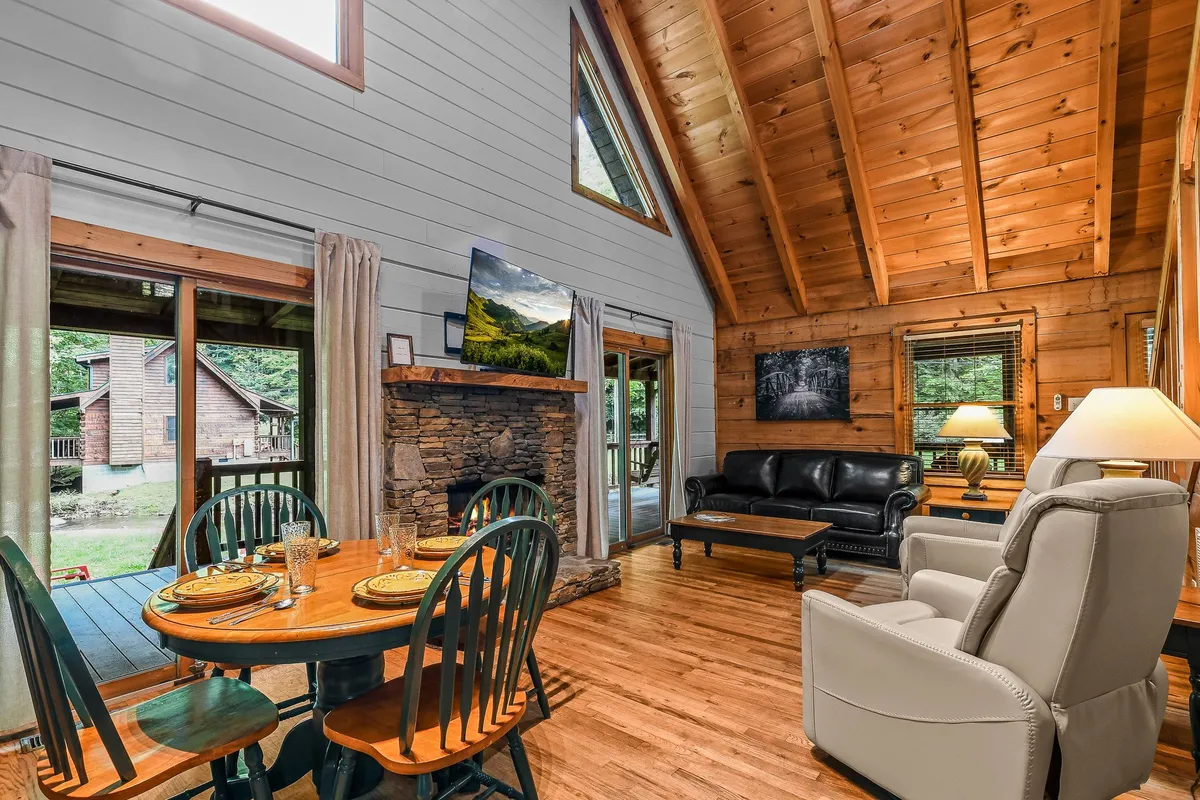 A spacious dining area with a large table set for a group, a cozy leather sofa, and a fireplace, all illuminated by generous natural light from sizable windows. The scene invites gatherings for meals, games, or relaxed conversations.