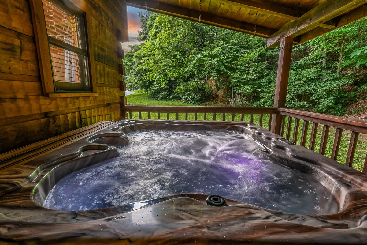 This exterior image showcases a wooden hot tub nestled amid lush greenery, with the wooden structure of the cabin visible in the background. The setting feels peaceful, inviting guests to relax after a day of outdoor adventures. The surrounding trees and foliage emphasize the cabin's immersion in nature.