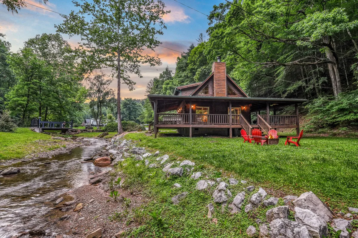 This exterior image showcases the cozy cabin in its natural setting, featuring red chairs placed near a stream and a visible fire pit area. The lush greenery and calm water establish a perfect environment for outdoor family activities like splashing in the creek, skipping stones, or simply relaxing by the water. The presence of the fire pit and surrounding seating hints at communal gatherings and storytelling under the stars.