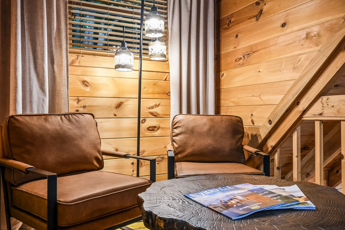 A welcoming nook featuring leather chairs around a wooden table, complemented by soft hanging light fixtures. The large windows provide a glimpse of the natural surroundings, blending indoor relaxation with an appreciation for the outdoors.