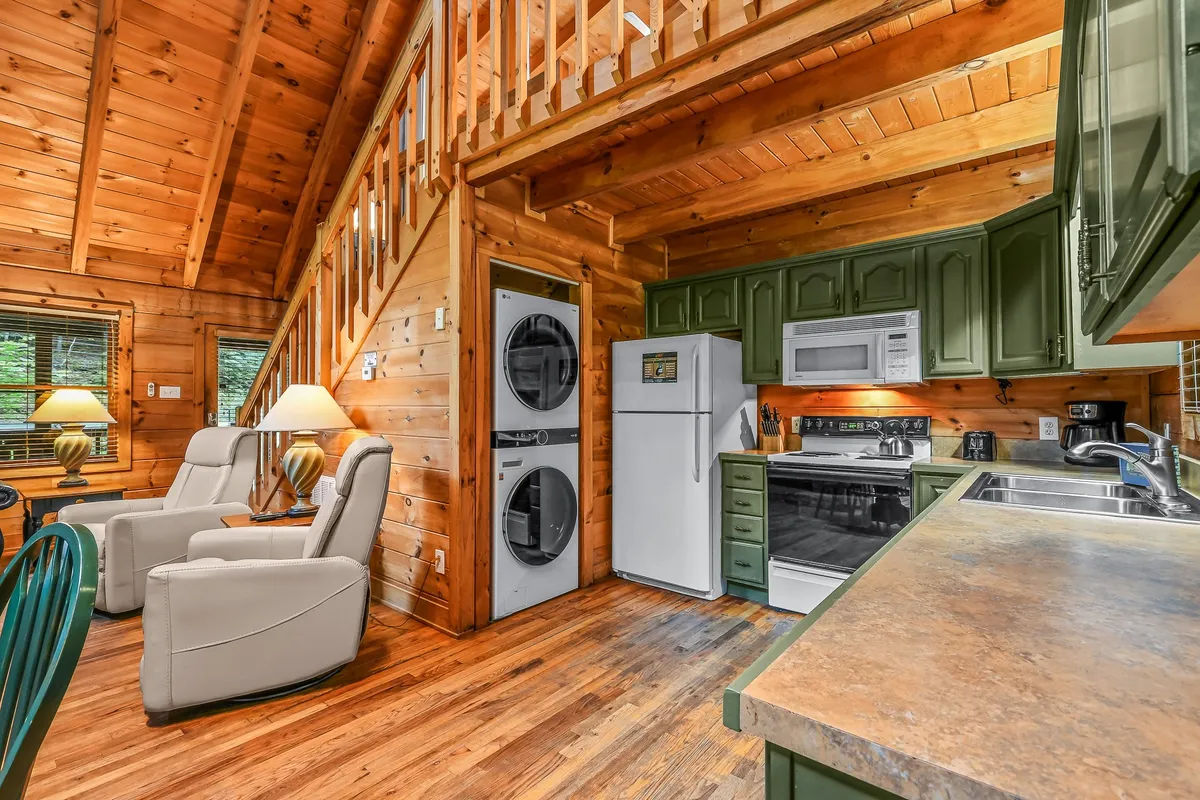 This image showcases a welcoming kitchen and dining area with wooden beams, green cabinetry, comfortable seating, and a dining table set near large windows. The space is bathed in natural light, emphasizing the inviting atmosphere and encouraging group gatherings for meals or conversation.